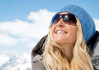 Woman in a heavy coat, sunglasses, and knit cap as she smiles in front of a mountanous background