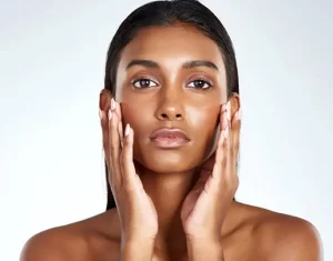 a dark skinned woman with her heair slicked back looks at the viewer while touching her face with both hands