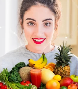 Woman looks over a collection of healthy food including pineapple, bannana, melon, tomatoes, apples, V8 in a glass, and other greens